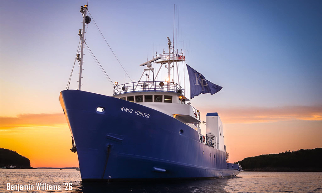 Blue research vessel “Kings Pointer” docked at sunset, with flag flying and calm water reflecting the sky.