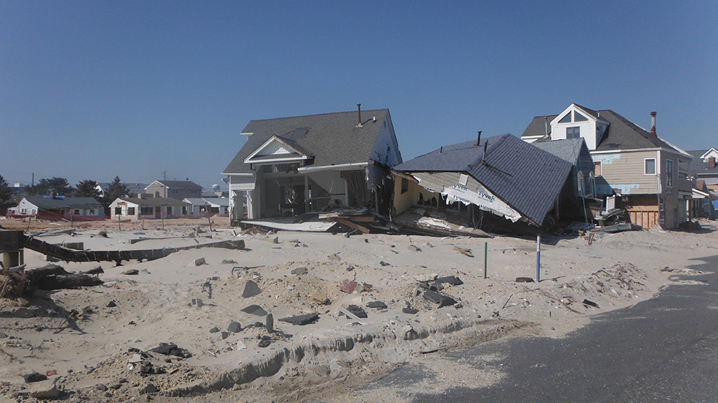 Two houses severely damaged, partially collapsed, and surrounded by sand and debris after a natural disaster.