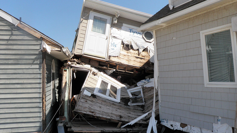 Two severely damaged houses with collapsed walls and debris between them under a clear blue sky.
